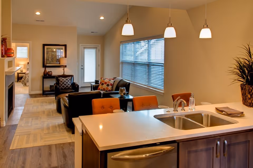Interior view of a modern assisted living facility showing a kitchen island with a double sink and three pendant lights above. In the background, there is a living area with black leather chairs, a sofa with colorful pillows, a side table, and a window with blinds. A hallway leads to another room with a bed visible.