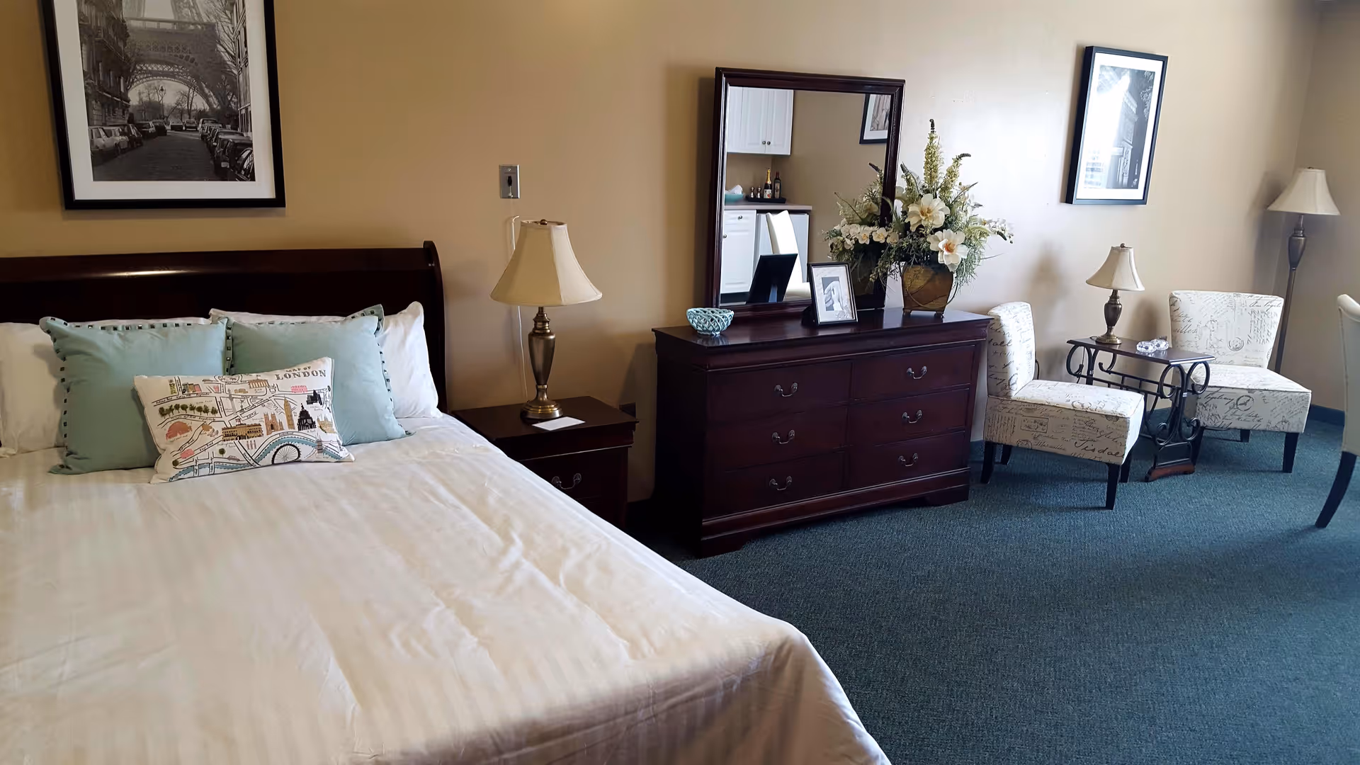 Bright furnished bedroom with a bed, nightstand and lamp, dresser with mirror and flowers, and two upholstered chairs by a small table.