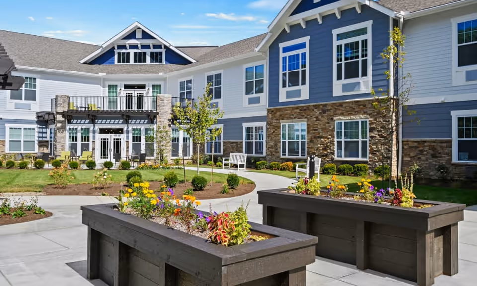 Outdoor courtyard area of The Ashton at Anderson senior living facility featuring raised garden beds with colorful flowers, a paved walkway, small trees, shrubs, benches, and a two-story building with blue and white siding and stone accents in the background under a clear blue sky.
