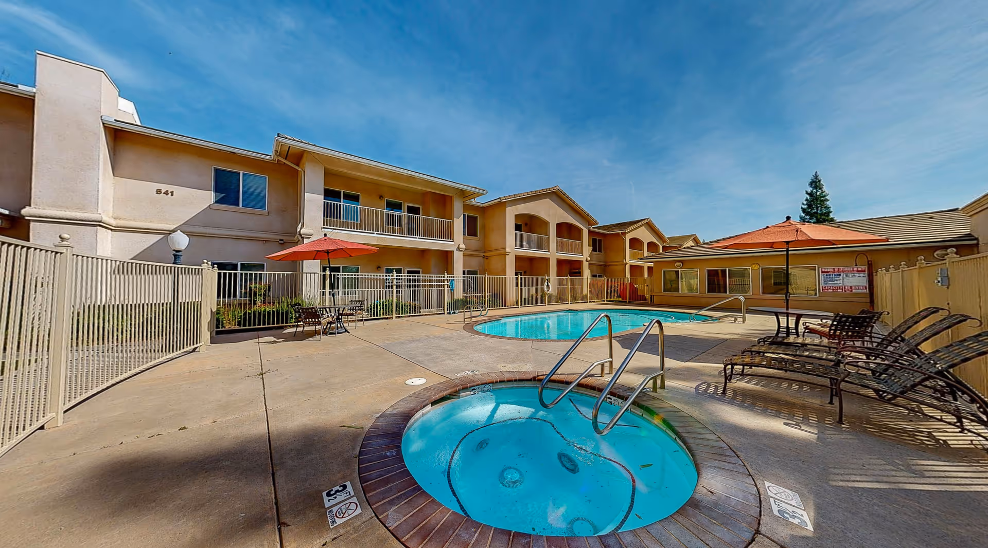 Outdoor pool area at Cogir of Cedar Creek featuring a swimming pool and a hot tub surrounded by a concrete deck. There are lounge chairs and tables with red umbrellas around the pool. The building with balconies and windows is visible in the background under a clear blue sky.