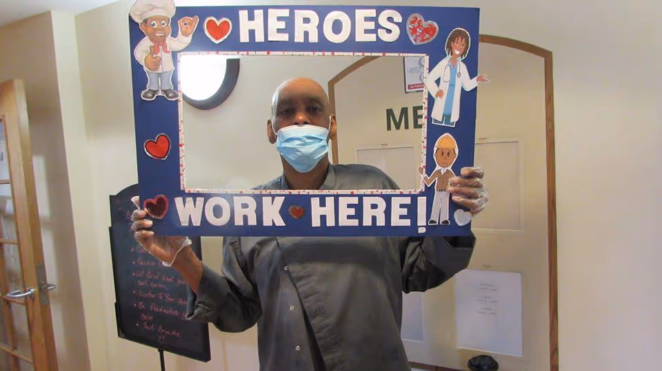 A person wearing a face mask and gloves holds a blue rectangular frame with cartoon characters and hearts. The frame has the text 'HEROES WORK HERE!' and the person is standing indoors in a hallway or common area.