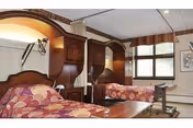 A nursing facility bedroom with two hospital-style beds, each with a floral patterned bedspread. The room has wooden headboards with built-in cabinets and medical equipment mounted on the wall. A large window with striped valance allows natural light into the room. There is a small table on wheels positioned near one of the beds.