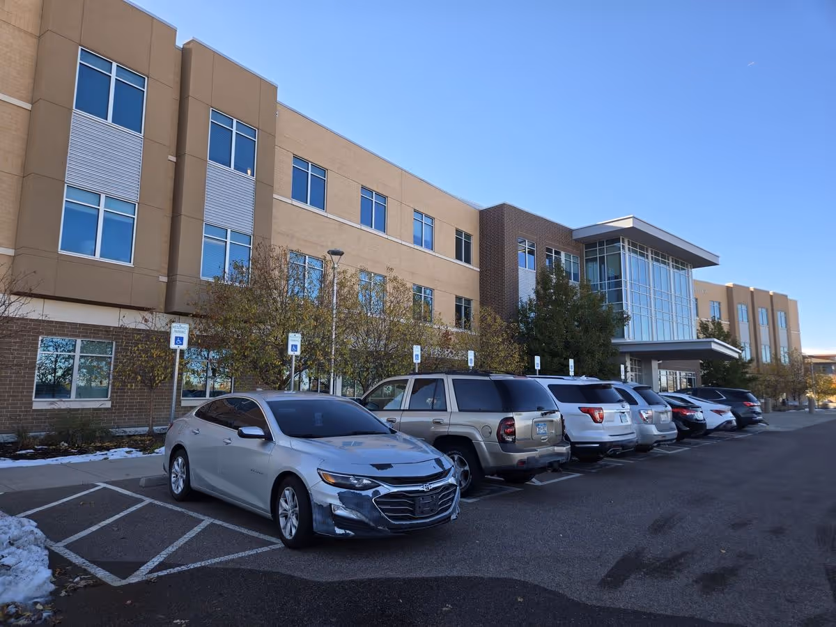 Exterior front of a three-story care facility with large windows and a row of parked cars in front under a clear blue sky.