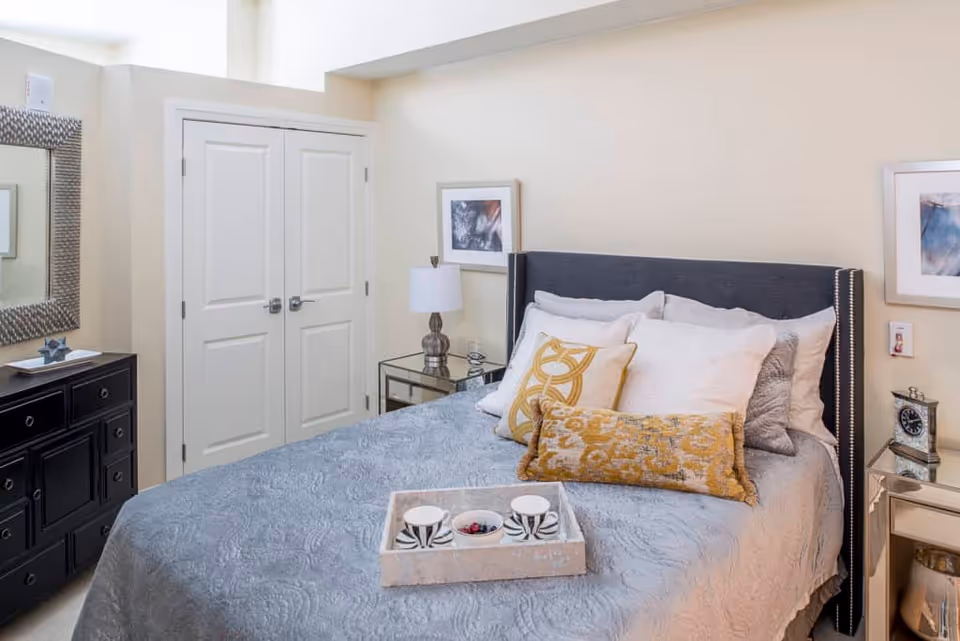 Bright, well-decorated bedroom with a made bed topped with pillows and a tray, bedside tables, lamp, and a dresser with mirror.