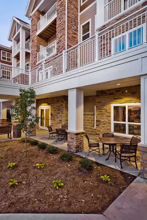 Outdoor patio area at Eagle Crest South with stone and wood exterior walls, several tables and chairs, a small tree, and landscaped garden beds under a covered walkway.