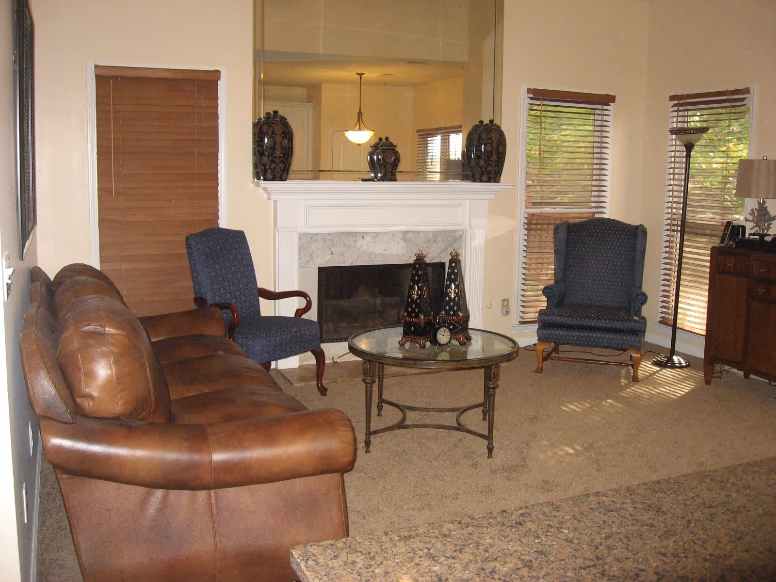 Cozy living room with a leather sofa, fireplace and mantle mirror, two armchairs, and a glass-top coffee table.
