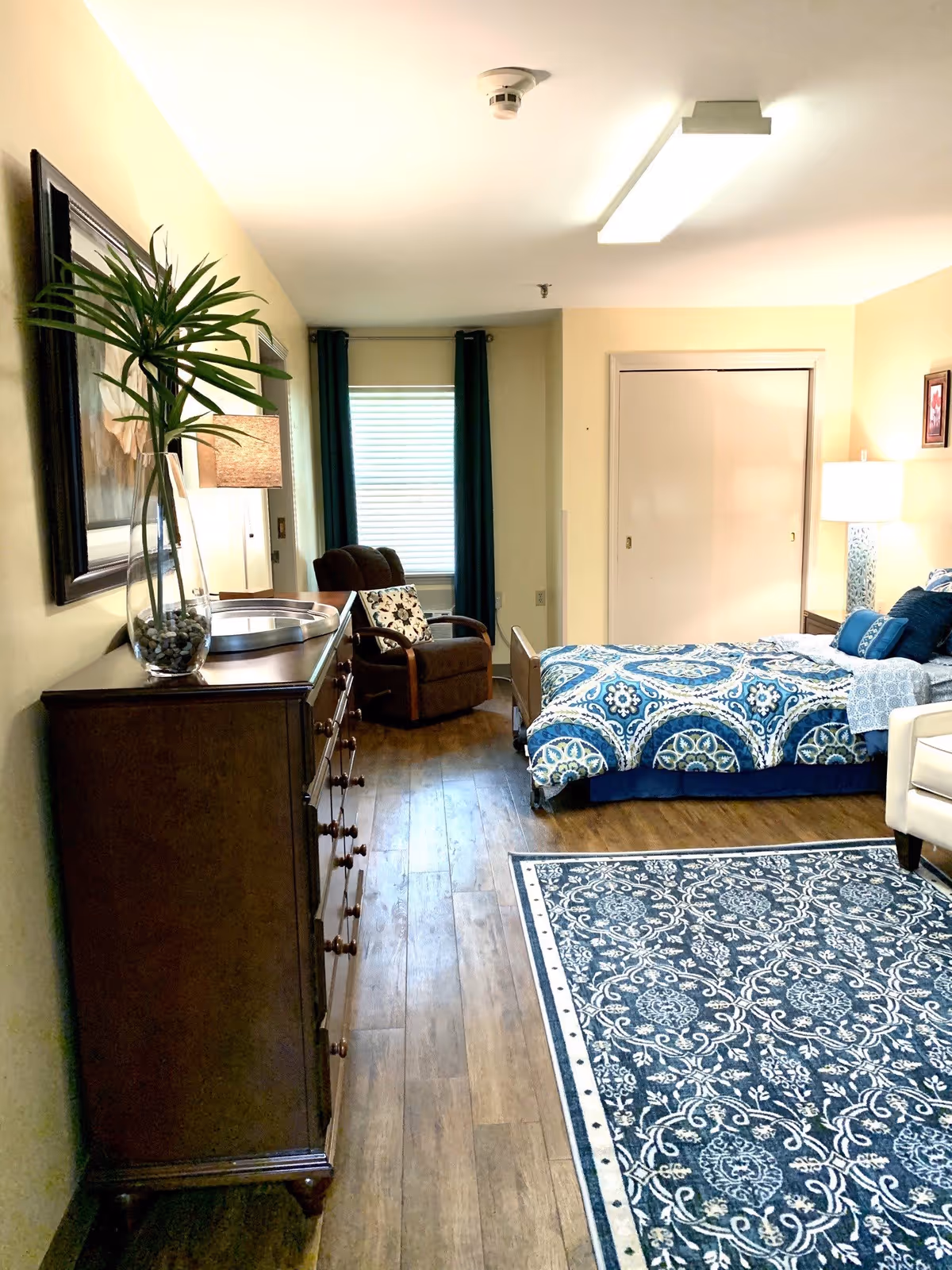Well-lit furnished bedroom with a bed dressed in blue patterned bedding, a wooden dresser, armchair by a window, and a patterned rug on wood floors.
