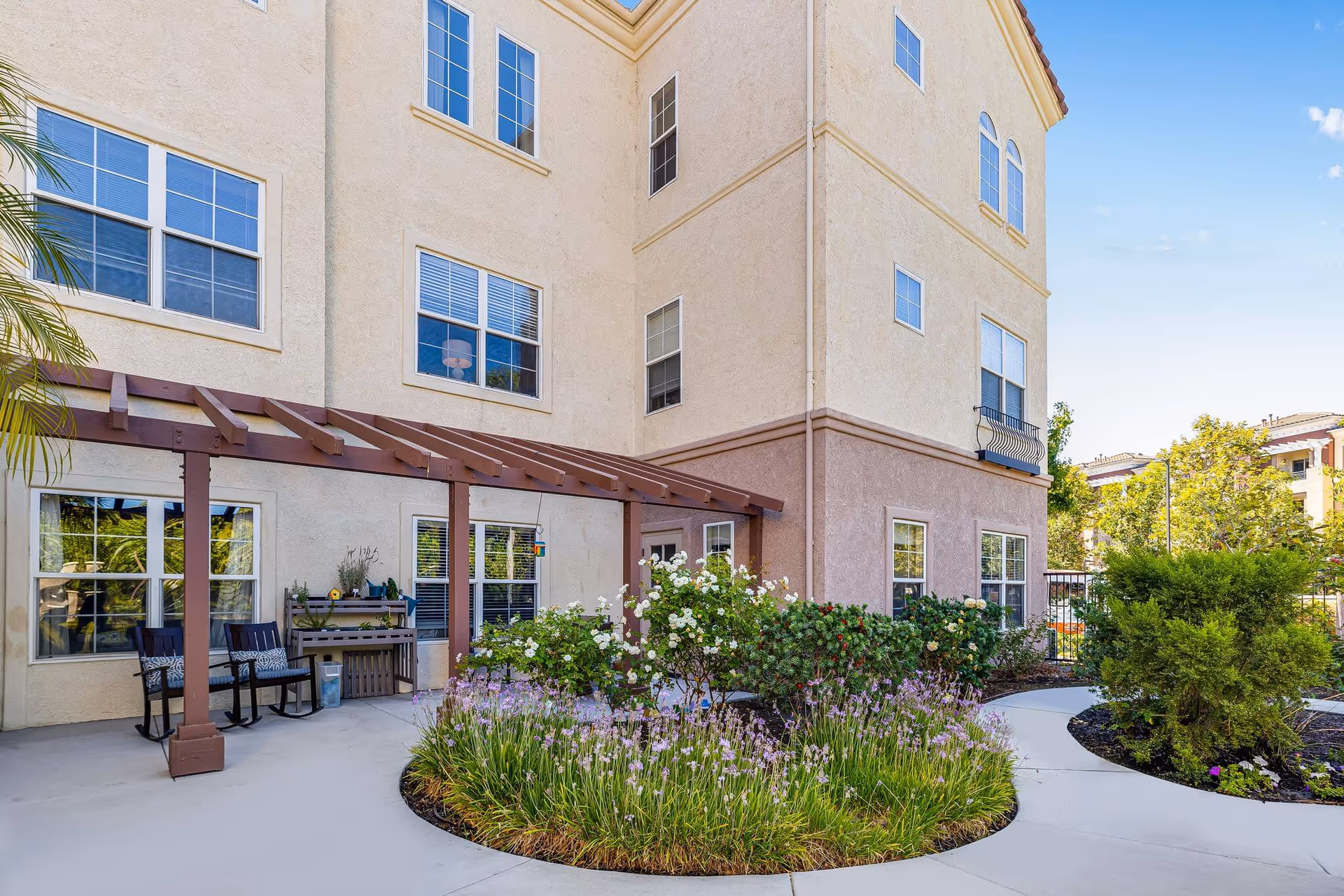 Outdoor patio area at a senior living facility with a beige multi-story building in the background. The patio has a wooden pergola, two black rocking chairs with cushions, a small table with plants, and a curved concrete walkway surrounded by landscaped flower beds with green shrubs and blooming flowers.