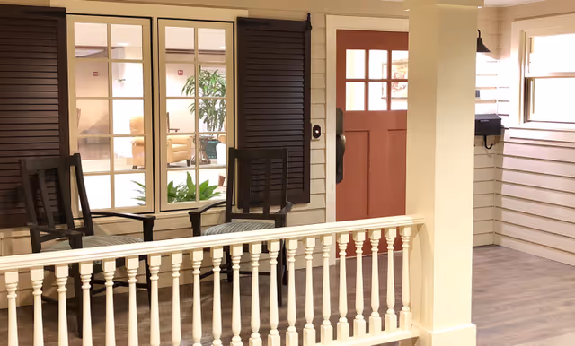 Interior mock porch area with two wooden chairs, white spindle railing, paneled walls, and a red front door.