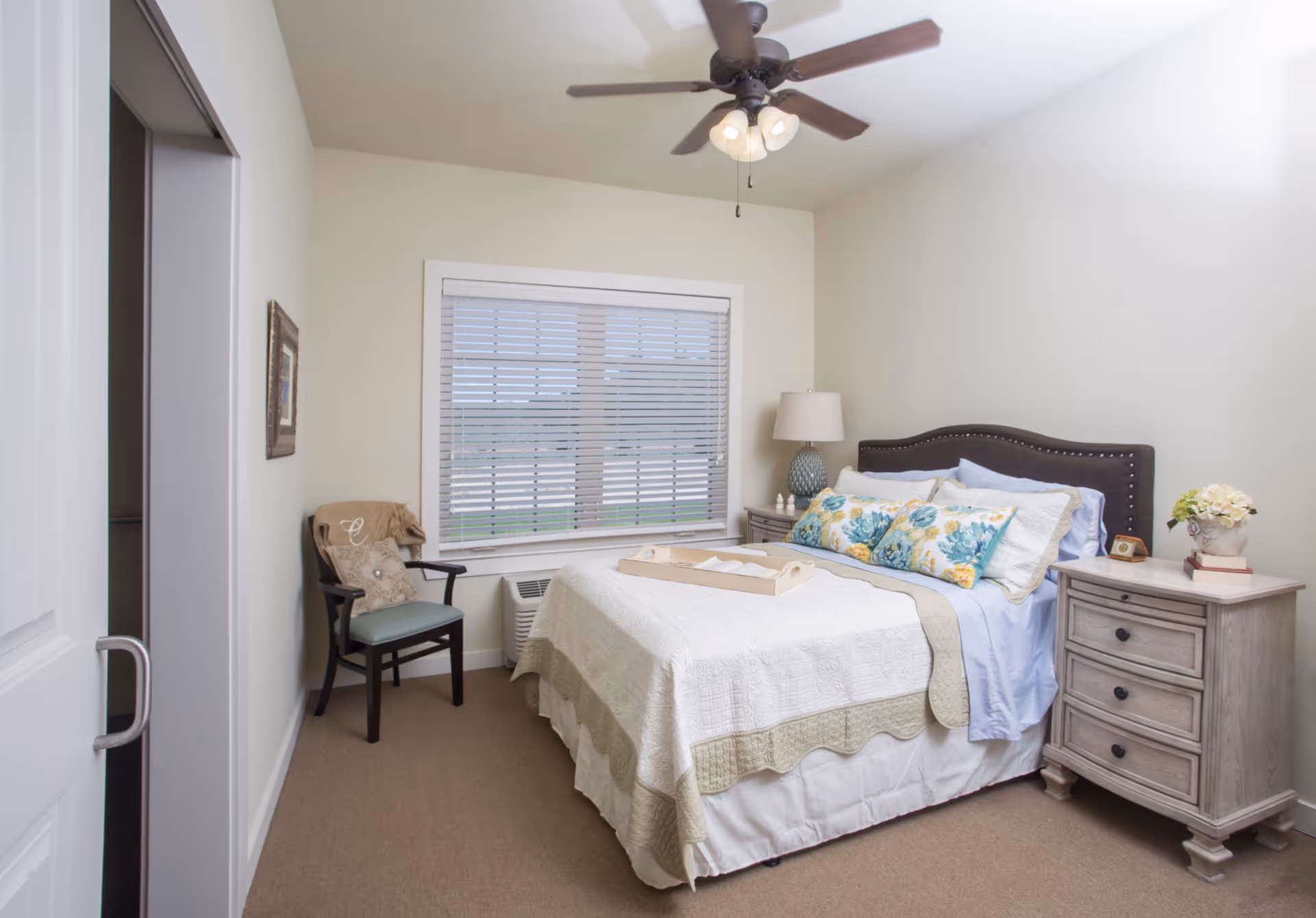 A cozy bedroom with a neatly made bed featuring a white and beige quilt, floral pillows, and a wooden tray on top. There is a wooden nightstand with a lamp and decorative items on the right side of the bed. A window with white blinds is centered on the wall behind the bed. A chair with a cushion and a blanket is placed near the window. The room has beige carpet and light-colored walls with a ceiling fan overhead.
