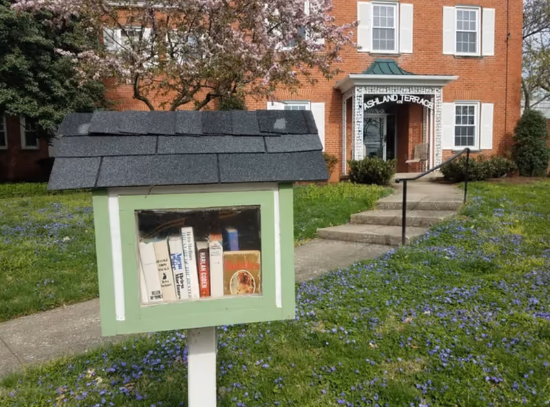 A small green and white wooden box with a shingled roof containing books, situated on a post in a grassy area with purple flowers. In the background, there is a brick building with white-framed windows and a white decorative entrance canopy labeled 'Ashland Terrace'. A concrete pathway with steps and a handrail leads to the entrance.
