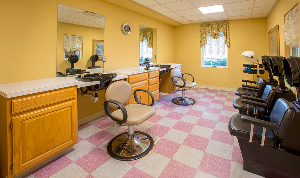 Interior view of a hair salon area with yellow walls, checkered pink and white floor tiles, two beige salon chairs in front of sinks and mirrors, and four black salon chairs with hair dryers along the right wall. There are two windows with decorative curtains and wooden cabinets under the sinks.