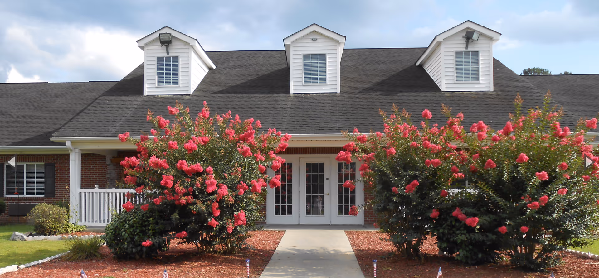 Front exterior view of a single-story brick building with a dark shingled roof and three dormer windows. The entrance features double glass doors and is flanked by large bushes with bright pink flowers. A concrete walkway leads up to the entrance, and the landscaping includes mulch and small American flags.