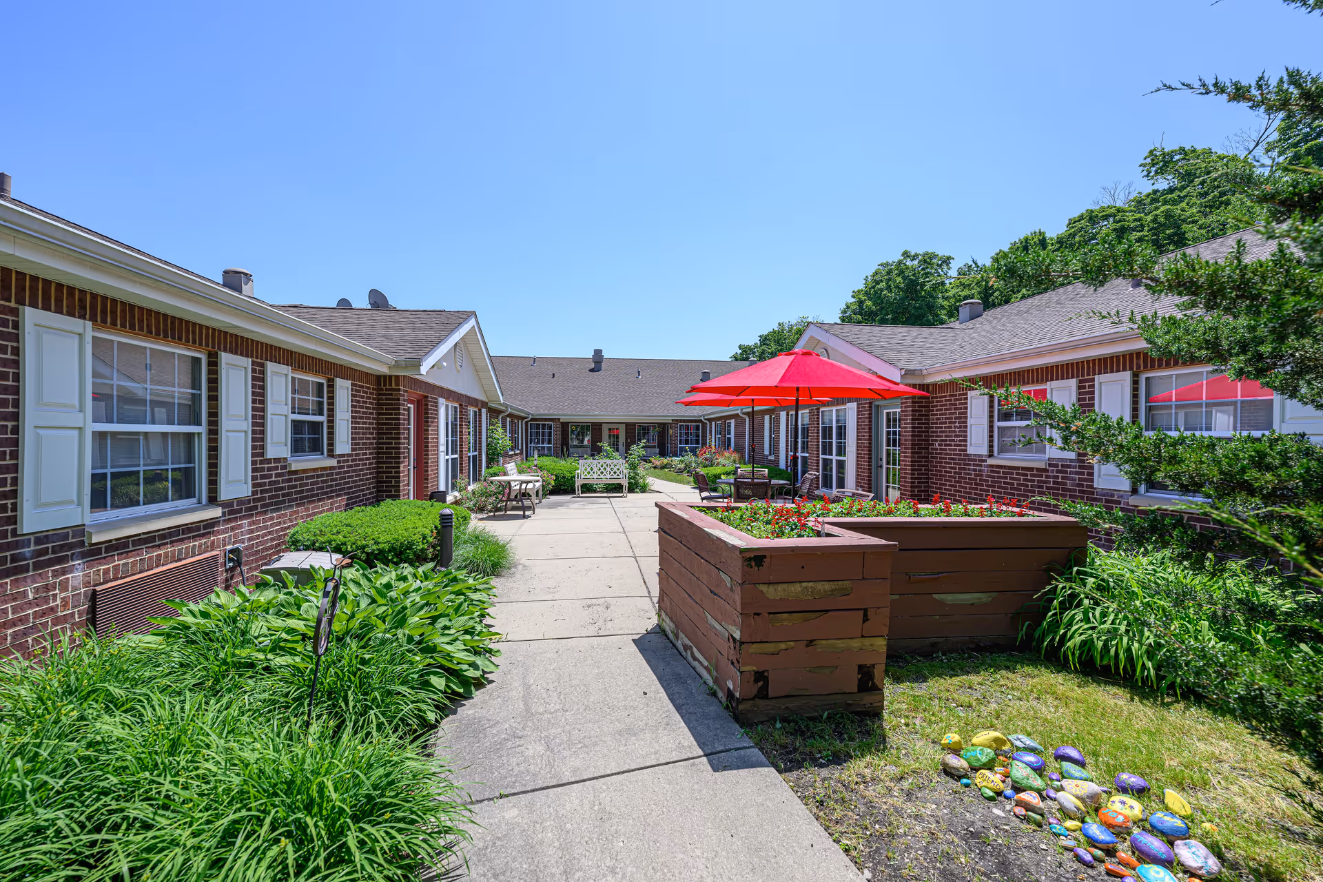 Outdoor courtyard area of Arden Courts - ProMedica Memory Care Community (Northbrook) featuring a paved walkway, green plants, raised garden beds with flowers, red patio umbrellas, benches, and brick buildings with white window shutters under a clear blue sky.