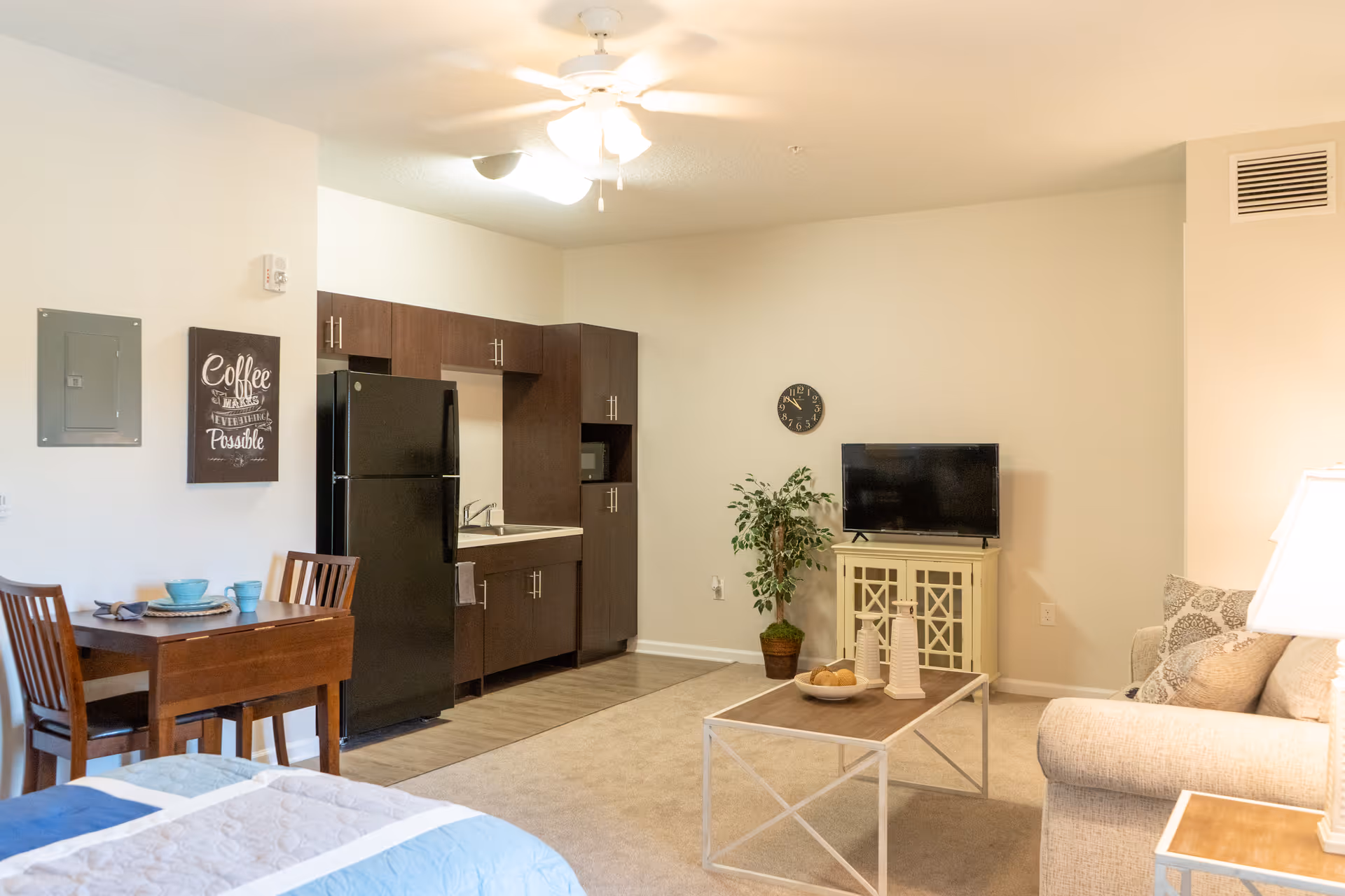 A cozy studio apartment interior featuring a small kitchen with dark cabinets and a black refrigerator, a wooden dining table with two chairs set with blue dishes, a beige sofa with patterned pillows, a coffee table with decorative items, a flat-screen TV on a white cabinet, a potted plant, and a ceiling fan with lights.