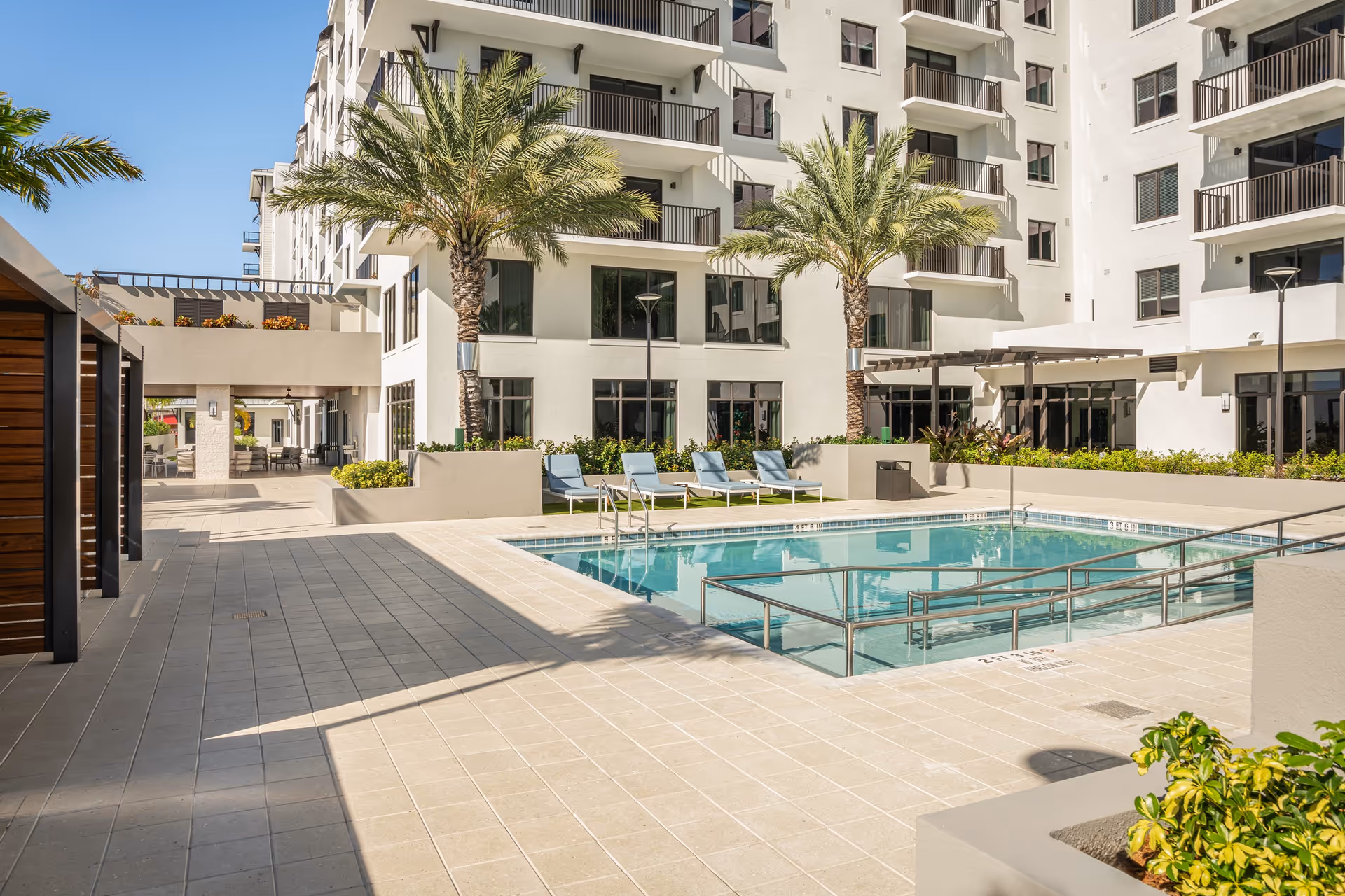 Outdoor pool area at Allegro – Fort Lauderdale, FL with lounge chairs, palm trees, and a modern multi-story building in the background under a clear blue sky.