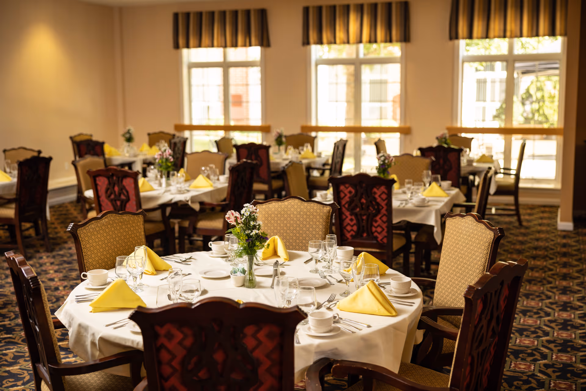 A dining room with multiple round tables set for a meal. Each table is covered with a white tablecloth and has yellow folded napkins, glassware, plates, and silverware. There are floral centerpieces on the tables. The room has large windows with striped valances allowing natural light to fill the space. The chairs have ornate wooden backs and upholstered seats.
