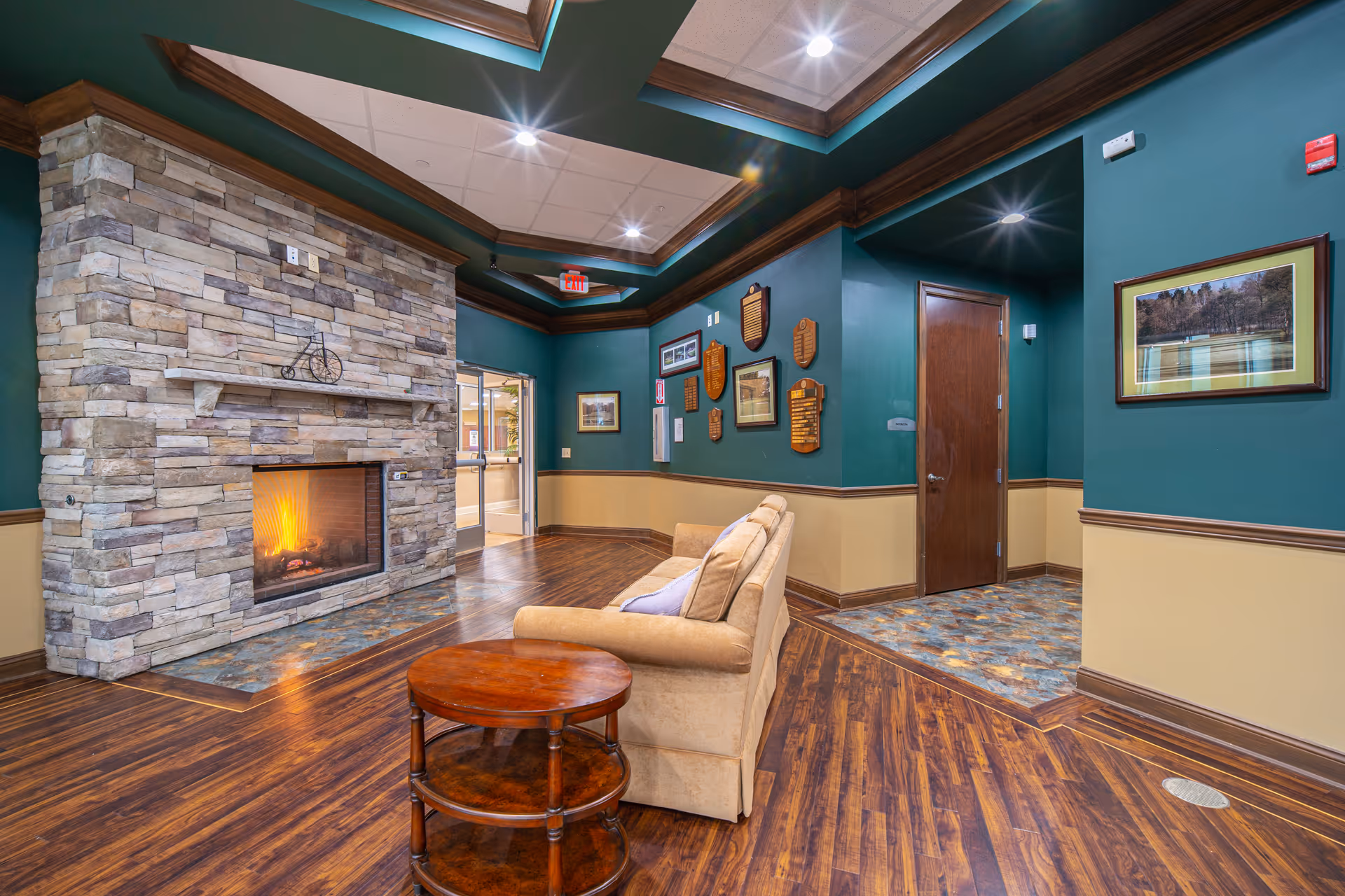Interior common lounge with a stone fireplace, beige sofa, round wooden table, and hardwood floors.