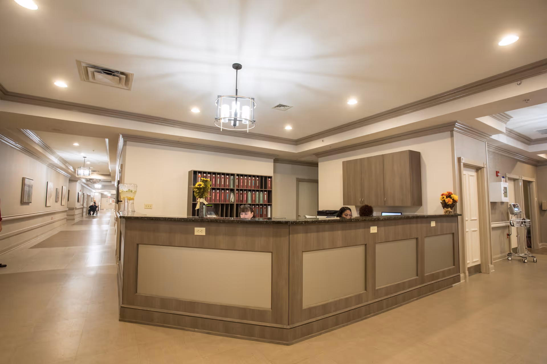 Reception desk area inside CareOne at Evesham Assisted Living facility with two staff members seated behind the desk. The desk has a granite countertop and wood paneling. Behind the desk are shelves with organized binders and a cabinet. The hallway extends to the left with framed pictures on the walls and ceiling lights.