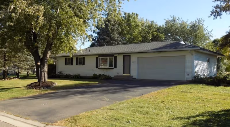 Single-story house with white siding, a two-car garage, and a driveway surrounded by green grass and trees under a clear blue sky.