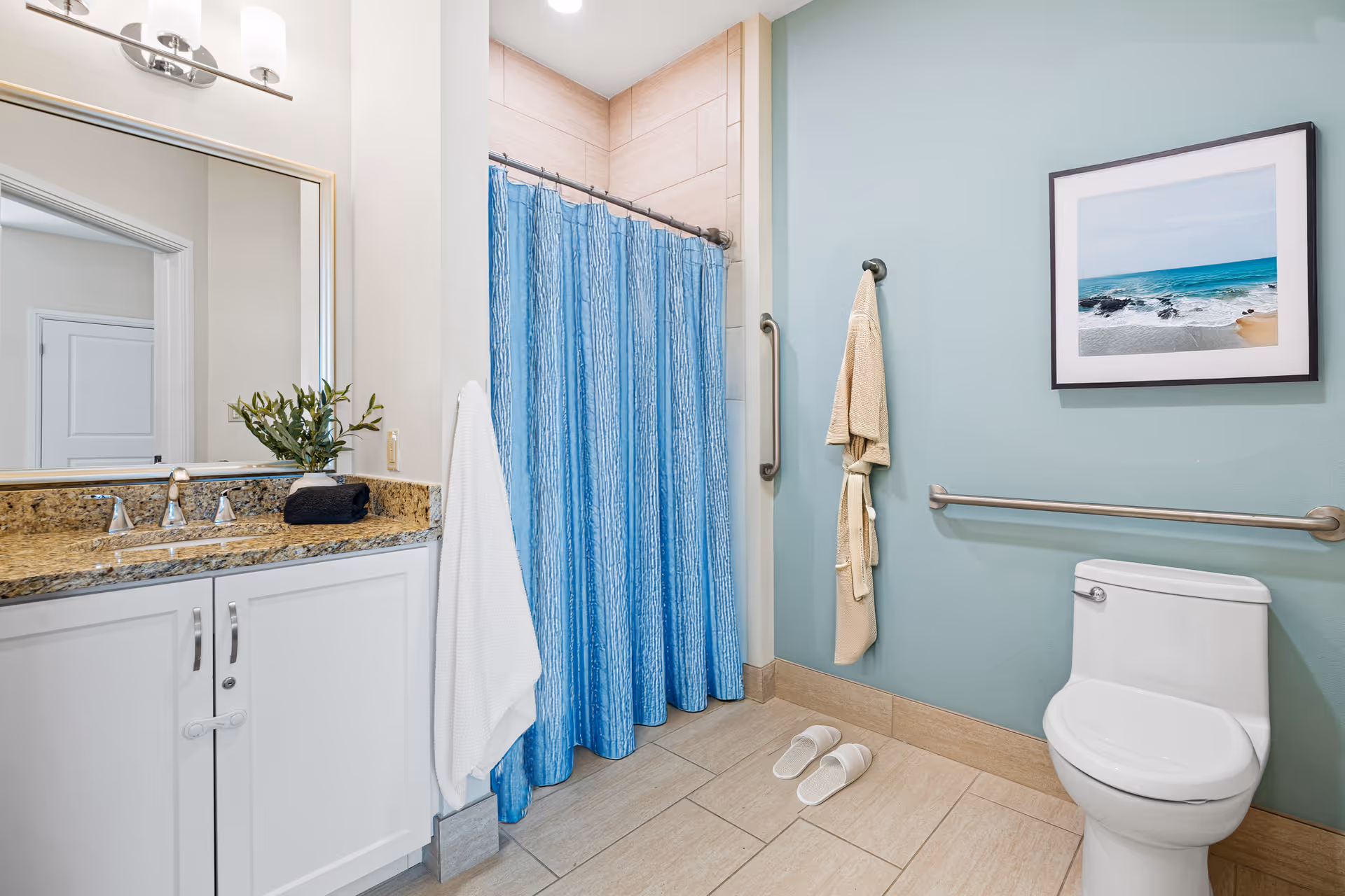 A clean and accessible bathroom featuring a white toilet with grab bars, a blue shower curtain, a beige bathrobe hanging on the wall, white slippers on the floor, a vanity with a granite countertop, a sink, a large mirror, and a framed picture of a beach scene on a light blue wall.