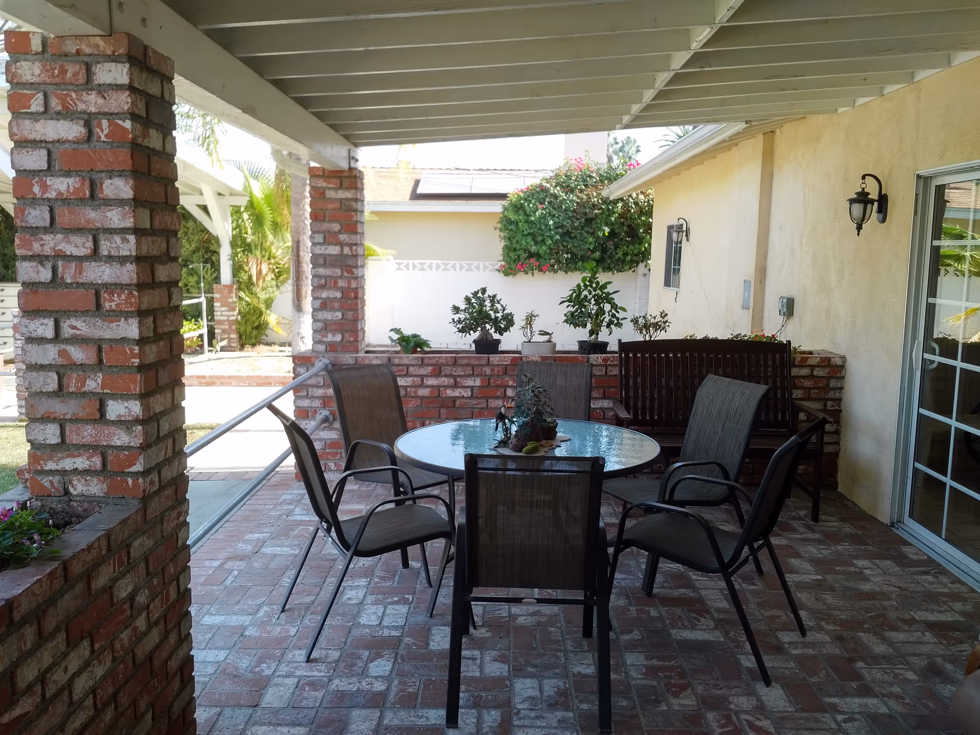 Covered outdoor patio area with a round glass table surrounded by five chairs and a wooden bench against a brick wall. Potted plants are placed on the brick wall and the patio floor is made of bricks. The patio is attached to a beige building with a sliding glass door and wall-mounted light fixtures.