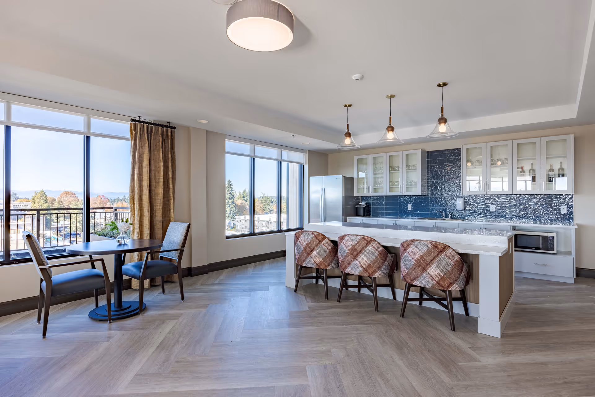 A modern kitchen and dining area with large windows offering a view outside. The kitchen features a long island with three patterned chairs, white cabinets with glass doors, a stainless steel refrigerator, and a microwave. There is also a small round table with two chairs near the windows, and pendant lights hanging above the island.