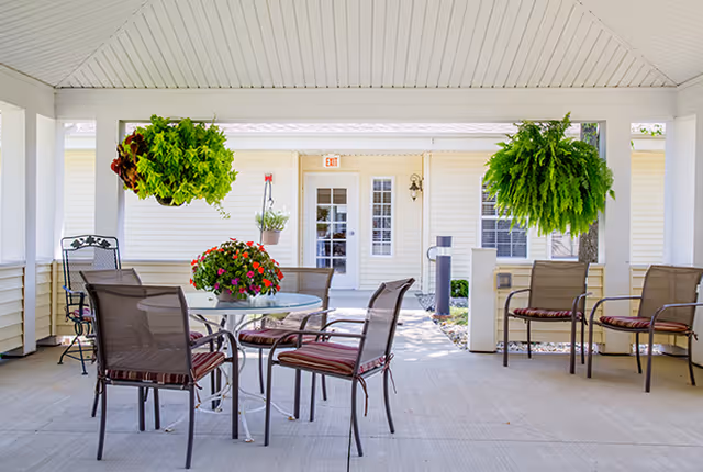 Covered outdoor patio area with a round glass table surrounded by six metal chairs with striped cushions. Two large hanging plants and a flower pot with red and pink flowers are visible. The background shows a white building with a door and windows.
