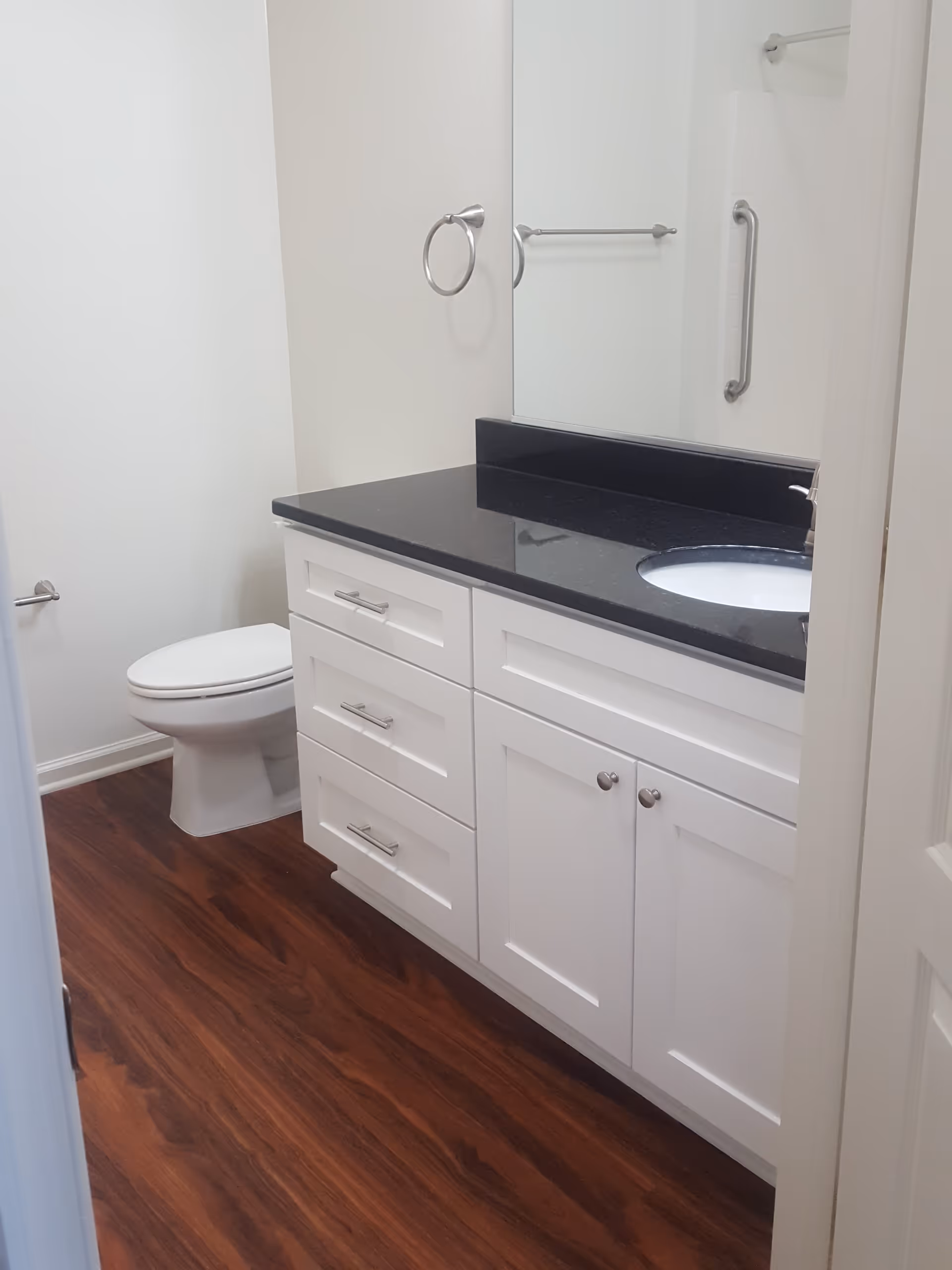 A clean bathroom with a white toilet, white cabinetry with silver handles, a black countertop with an inset sink, a large mirror above the sink, a towel ring, and a towel bar with a grab bar in the shower area. The floor has a wood-like finish.