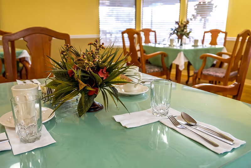 A dining table set with a glass, cup, saucer, and silverware on a napkin, with a floral centerpiece. In the background, there are more tables with green tablecloths and wooden chairs, and windows with blinds letting in natural light.