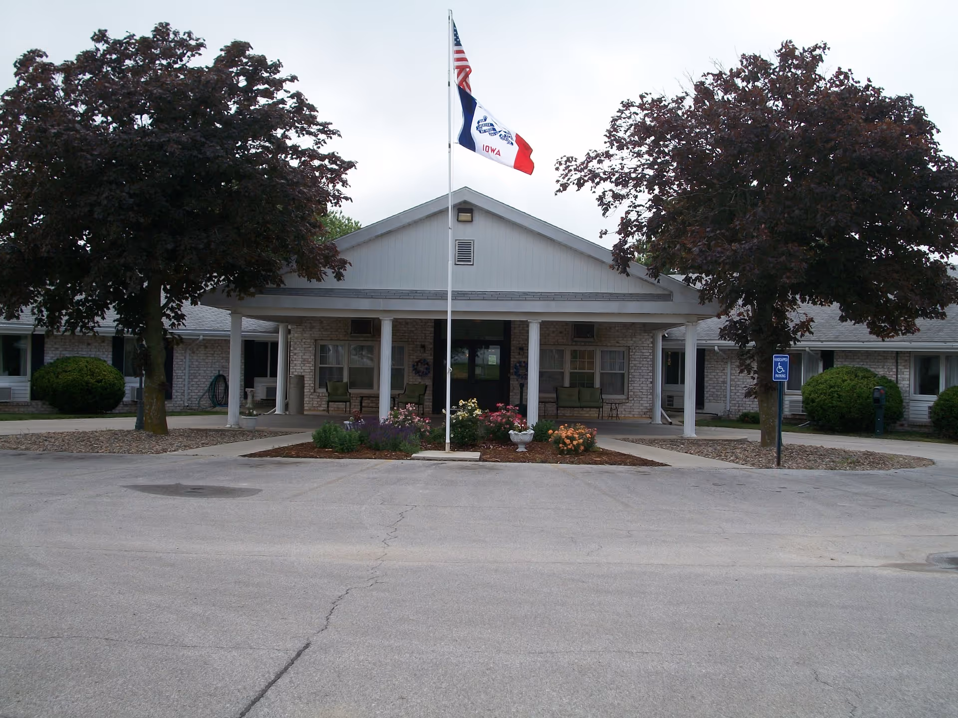 Front entrance of a single-story care facility with a flagpole, two trees, flower beds, and a paved driveway.