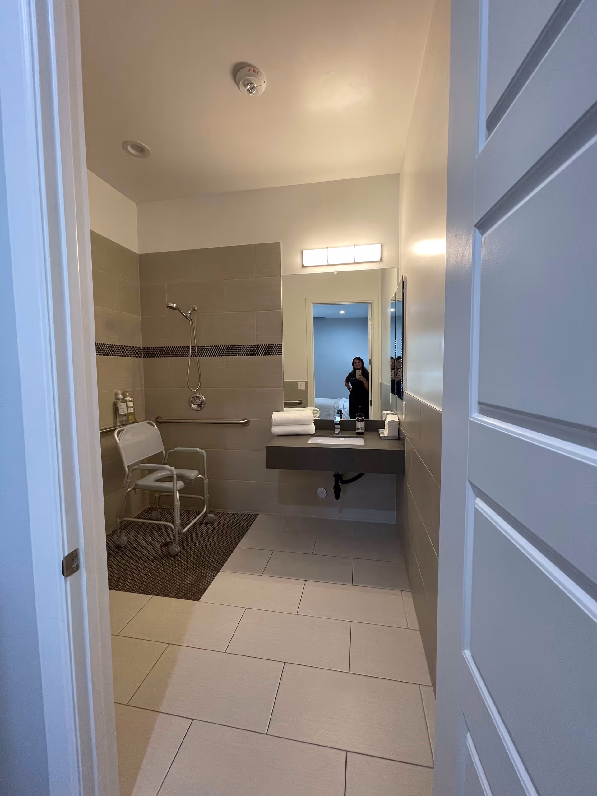 View into a modern bathroom with beige tiled walls and floor. There is a shower area with a handheld showerhead and a white shower chair. A dark gray floating sink countertop holds folded white towels, soap dispenser, and tissue box. A large mirror above the sink reflects a person taking the photo from an adjacent room. The bathroom door is partially open.