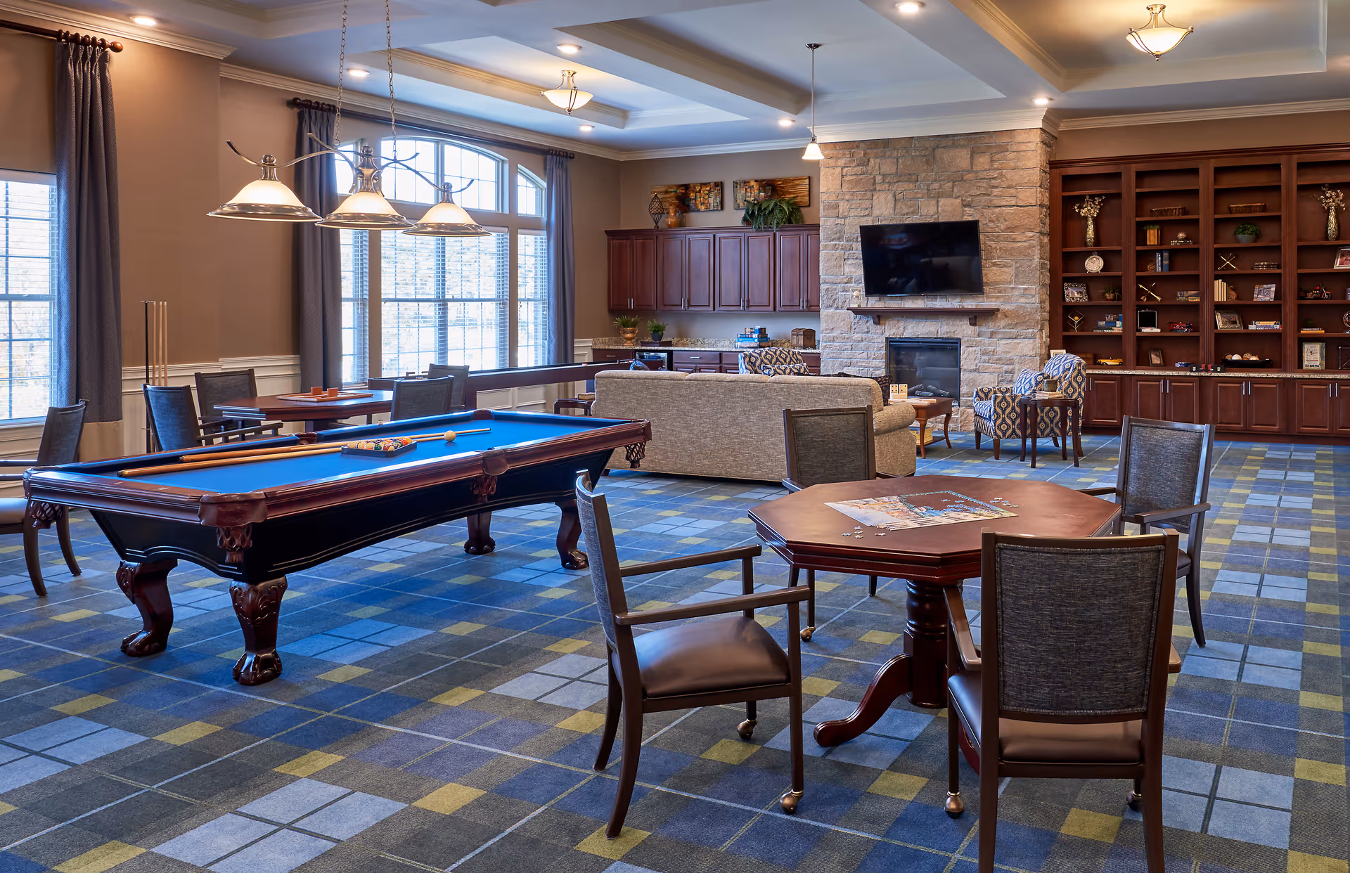 A spacious common area with a pool table in the center, surrounded by chairs and tables. The room features large windows with curtains, a stone fireplace with a mounted flat-screen TV above it, and built-in wooden bookshelves filled with decorative items. There is a comfortable seating area with a sofa and armchairs near the fireplace. The floor is covered with a patterned carpet in shades of blue, gray, and yellow.