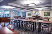 Spacious dining/activity room with rows of tables and chairs, large windows, a buffet sideboard, and framed artwork on the walls.