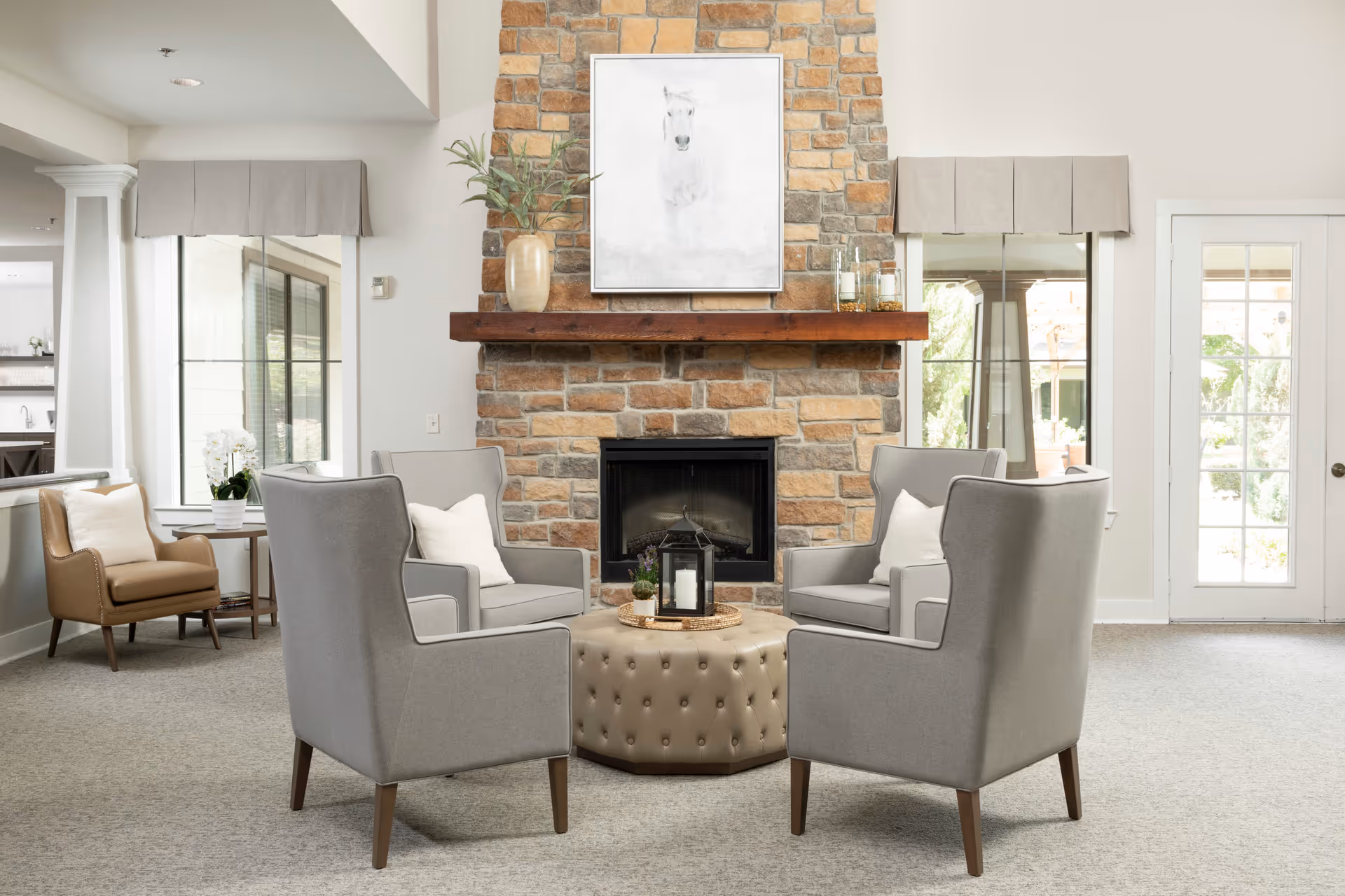 A bright sitting room with four gray armchairs arranged around a tufted ottoman in front of a stone fireplace.