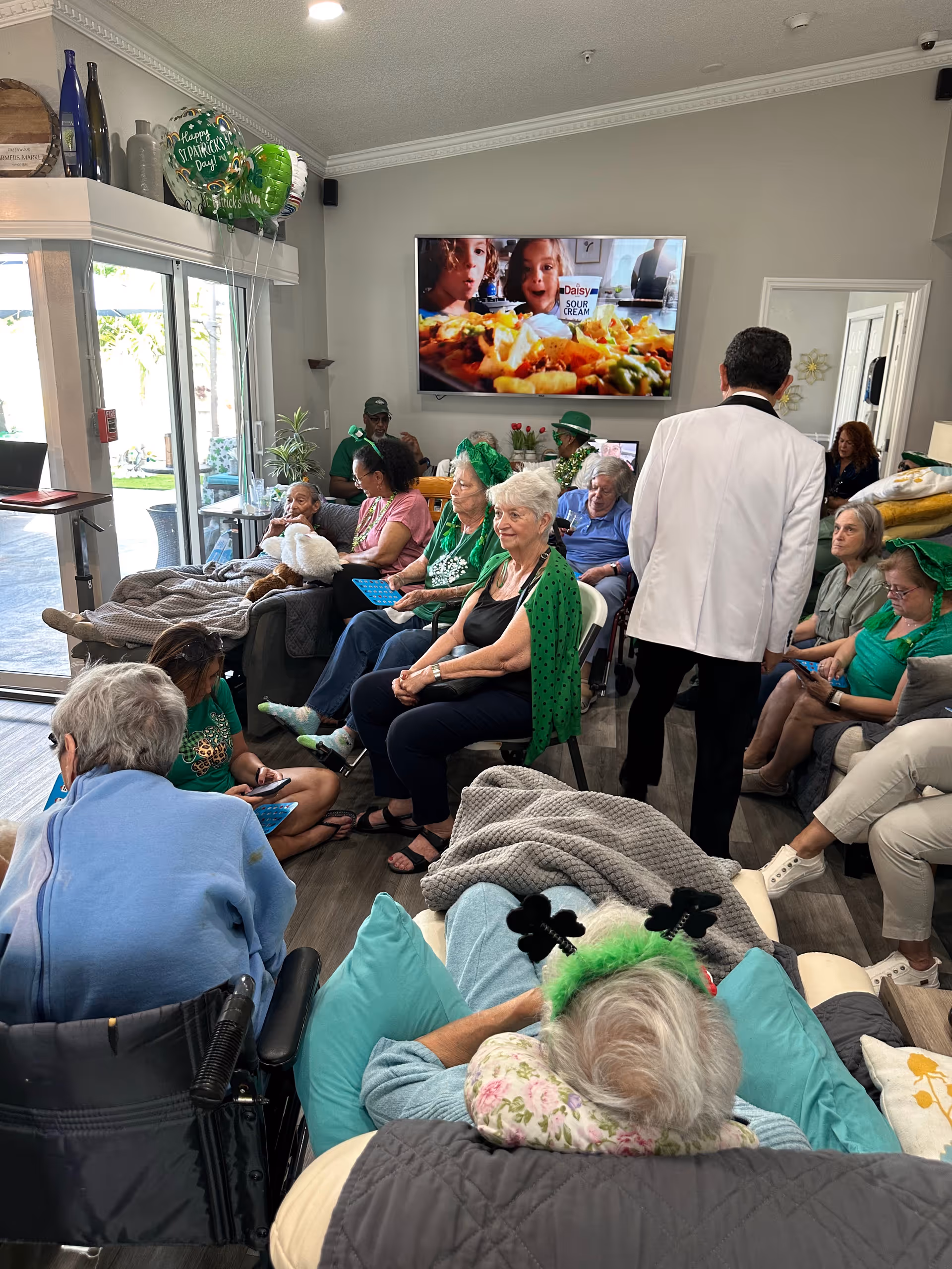 A group of elderly residents seated in a communal living room decorated for St. Patrick's Day with a large TV on the wall.
