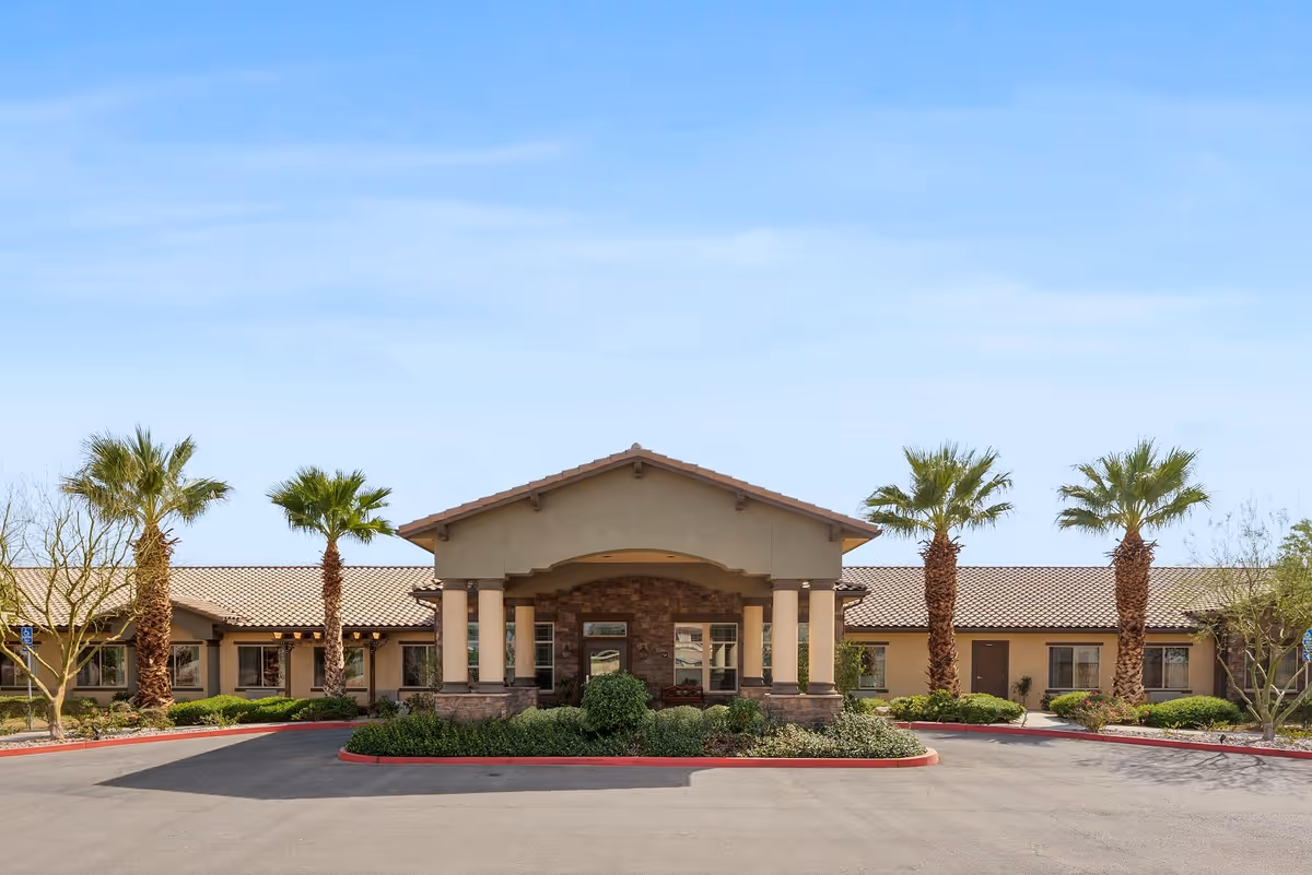 Front exterior view of Caleo Bay Assisted Living and Memory Care facility with a covered entrance, beige walls, stone accents, and palm trees lining the driveway under a clear blue sky.