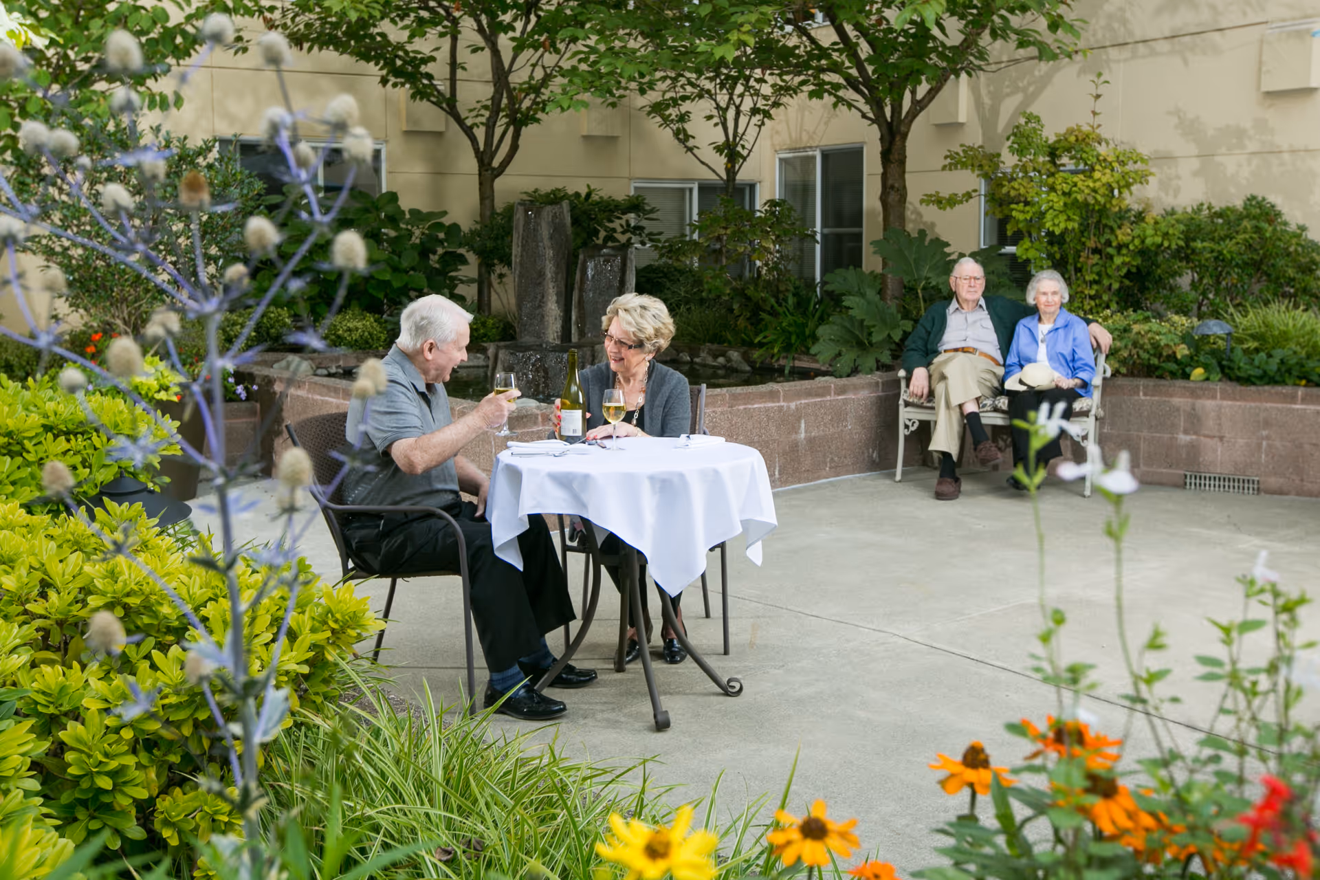 Two older adults share drinks at a small table in a landscaped outdoor courtyard while another couple sits on a bench in the background.