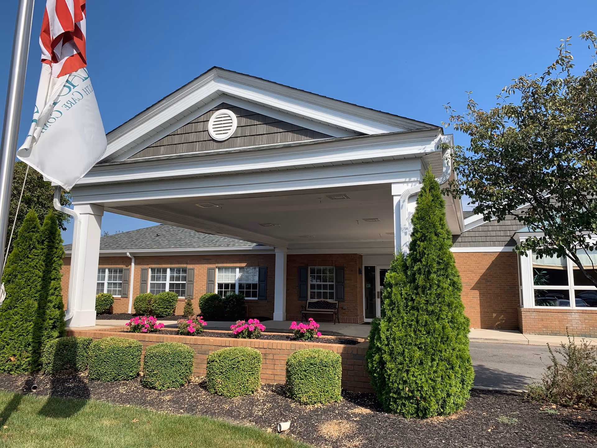Covered porte-cochère entrance of a brick senior living building with columns, landscaping, and flags.