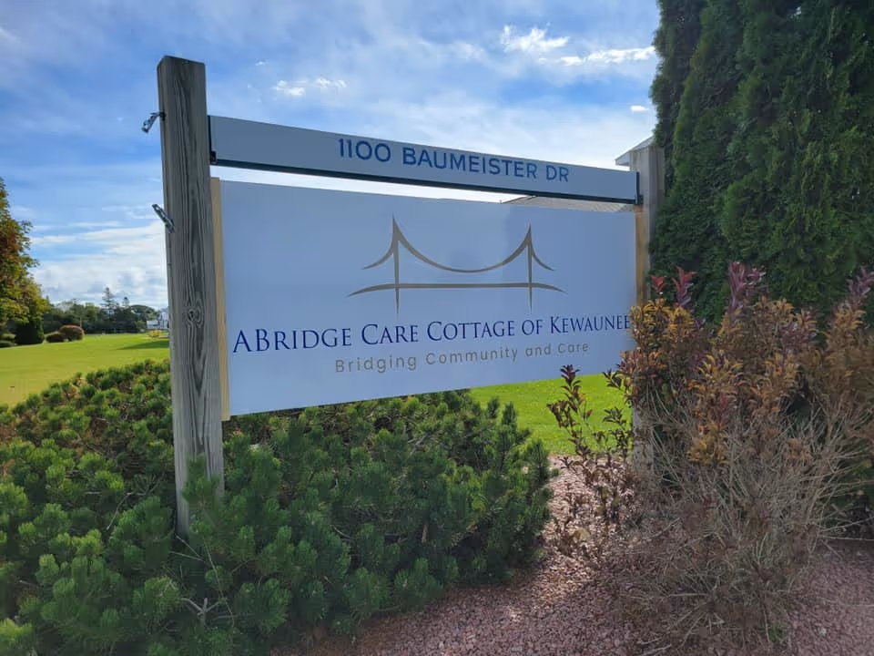 Outdoor sign for ABridge Care Cottage of Kewaunee located at 1100 Baumeister Dr, surrounded by green bushes and trees under a partly cloudy sky.