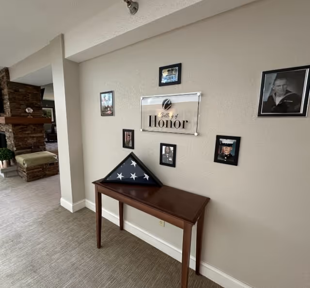 Indoor hallway wall with a wooden table holding a folded American flag beneath a 'Gallery of Honor' plaque and framed photos.