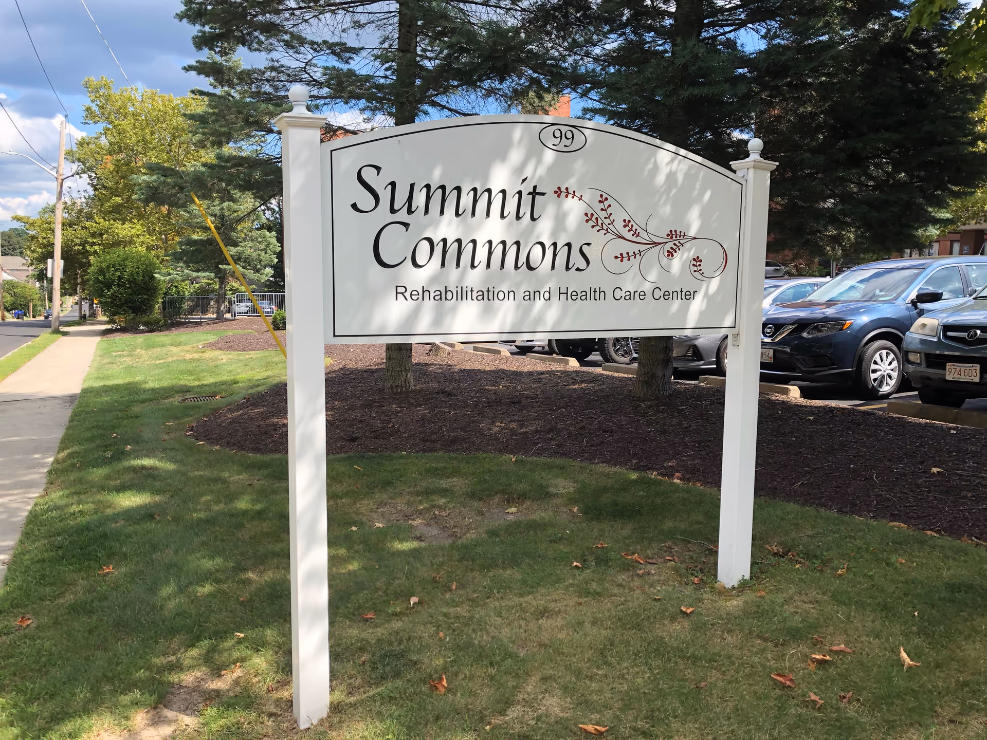 A white sign with decorative red accents reading 'Summit Commons Rehabilitation and Health Care Center' is displayed on a grassy area next to a sidewalk and a parking lot with several cars. Trees and a partly cloudy sky are visible in the background.