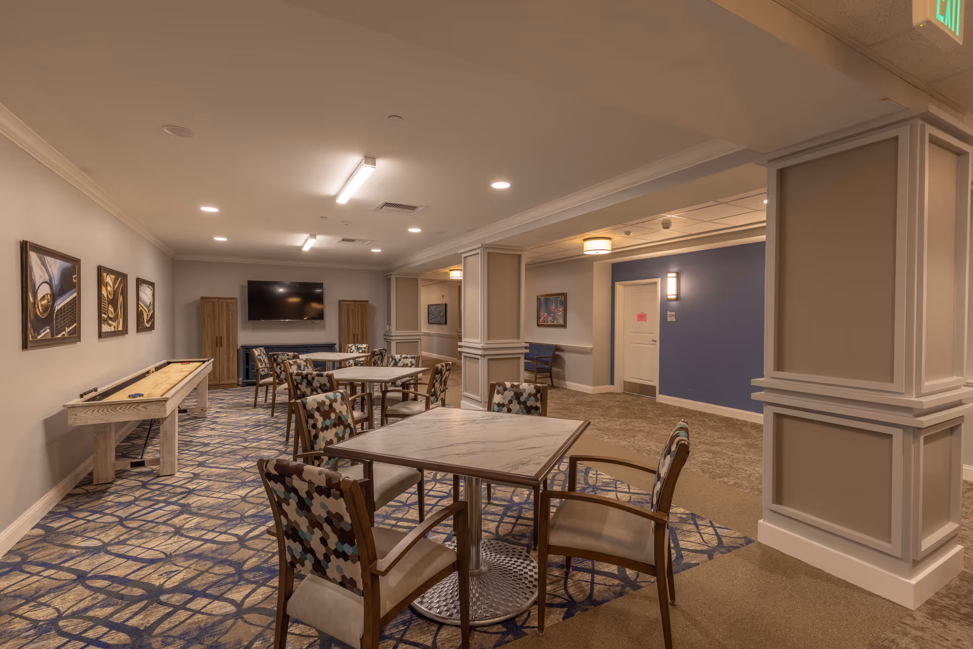 A well-lit common area in a senior living facility featuring several square tables with patterned chairs around them. The room has a carpeted floor with a blue and beige geometric pattern, beige walls, and decorative columns. On one side, there is a shuffleboard table and framed pictures on the wall. A flat-screen TV is mounted on the far wall between two wooden cabinets. The hallway adjacent to the room has a blue accent wall with a door and a bench underneath a framed picture.