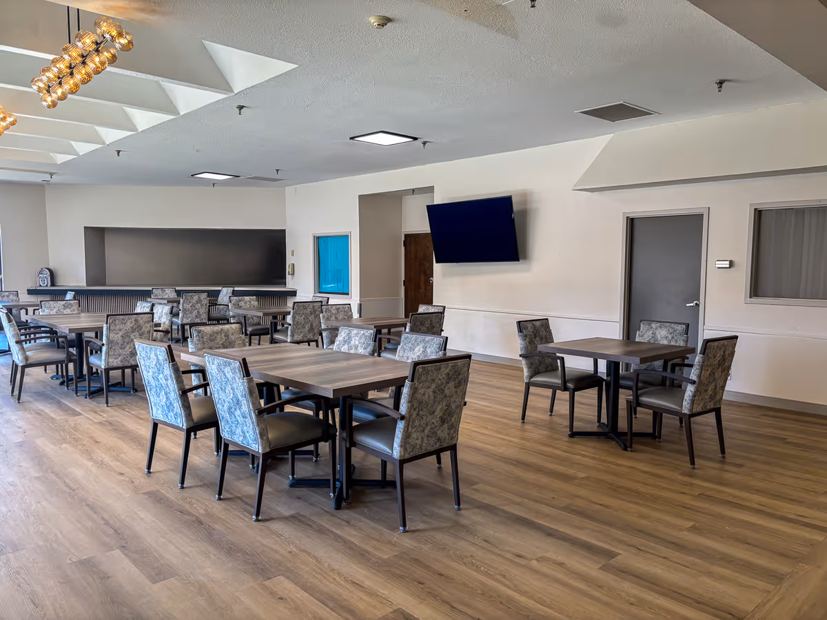 Spacious senior dining room with multiple tables and upholstered chairs, a wall-mounted TV, and wood-look flooring.