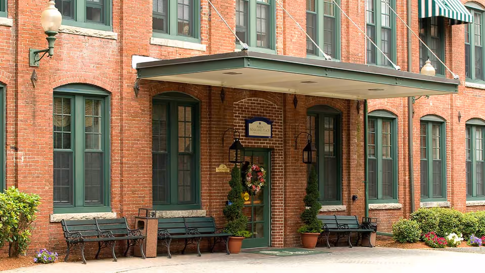 Exterior view of a brick building with green-framed windows and a green door decorated with a floral wreath. There are benches and potted plants near the entrance, which is covered by a green awning. A sign above the door reads 'Atria Marland Place'.