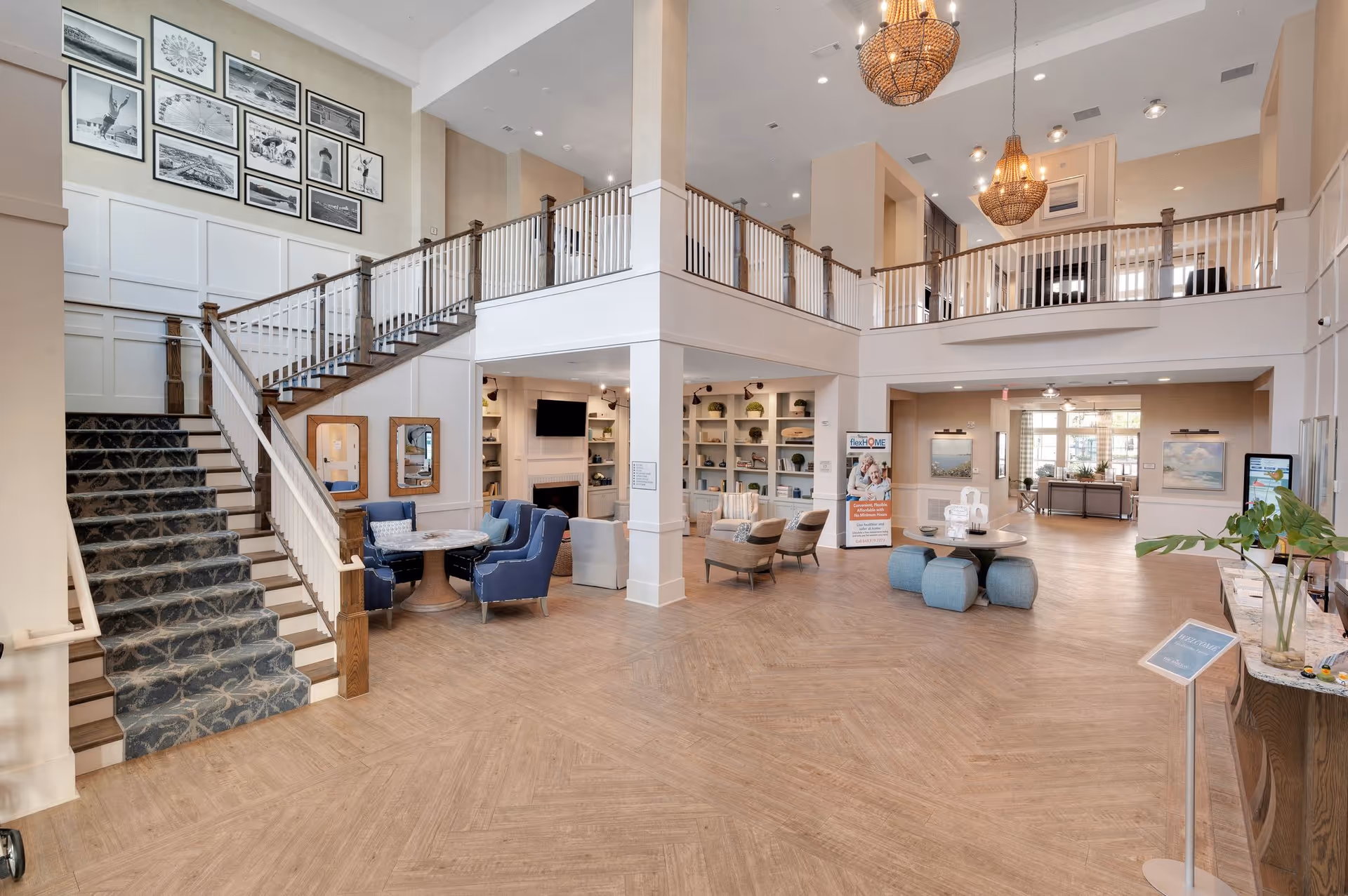 Spacious and elegant senior living facility lobby with a large staircase on the left, blue and beige seating areas, decorative wall art, chandeliers hanging from the high ceiling, and open spaces with wooden flooring.