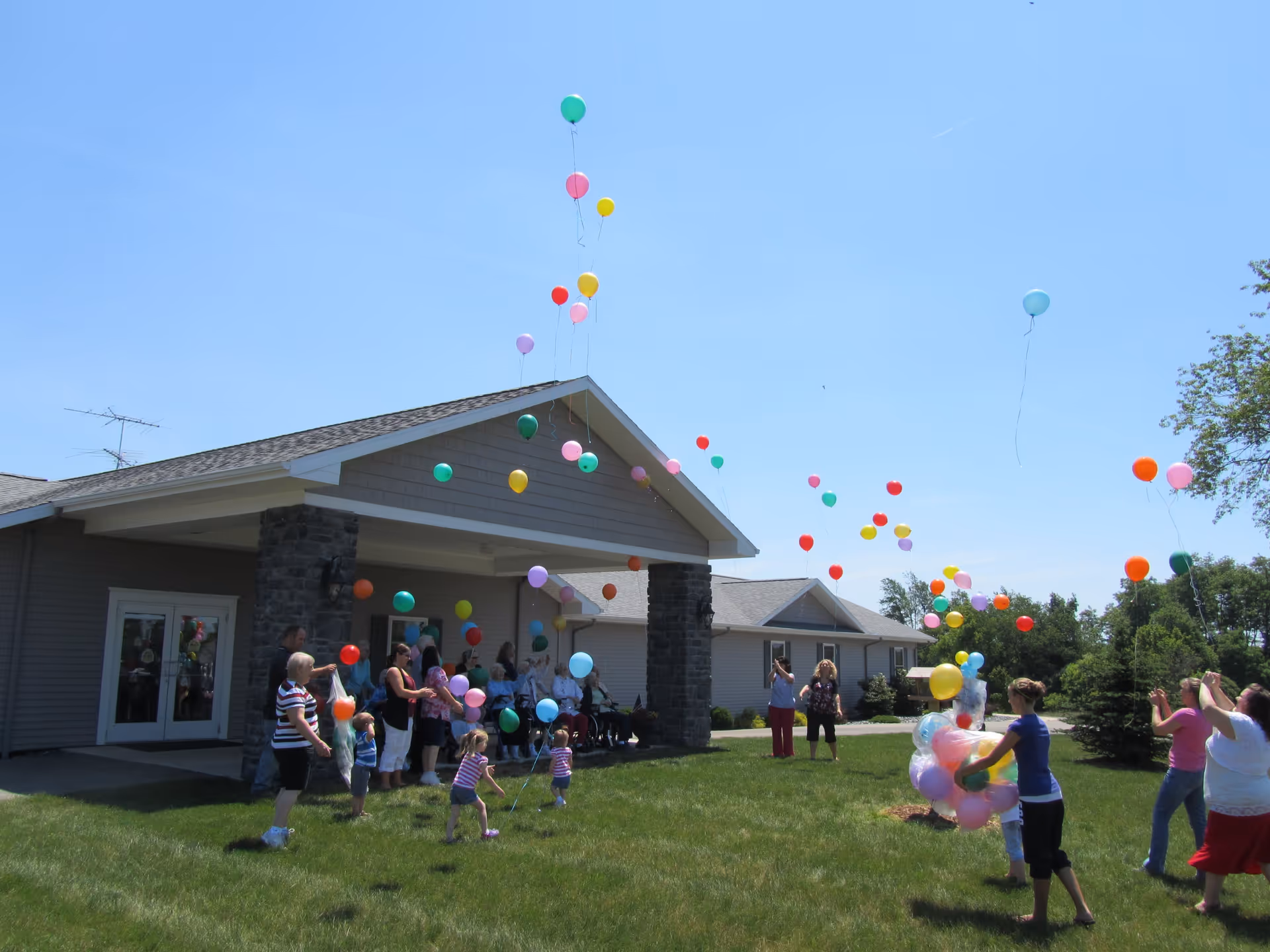 A group of people, including children and adults, are outside a single-story building with stone pillars and a covered entrance. They are releasing colorful balloons into a clear blue sky on a sunny day. The scene is lively with green grass and some trees in the background.