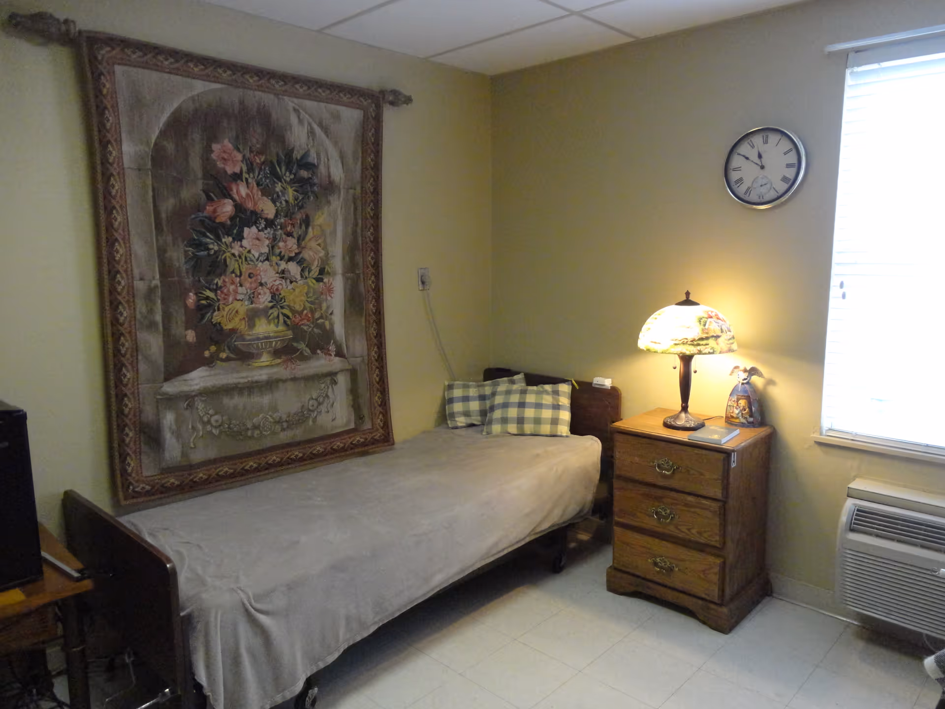 A small bedroom with a single bed covered with a gray blanket and two checkered pillows. Above the bed hangs a large tapestry featuring a floral arrangement. Next to the bed is a wooden nightstand with three drawers, a decorative lamp, and a small figurine. A clock is mounted on the wall above the nightstand, and a window with blinds is partially visible. An air conditioning unit is installed below the window.
