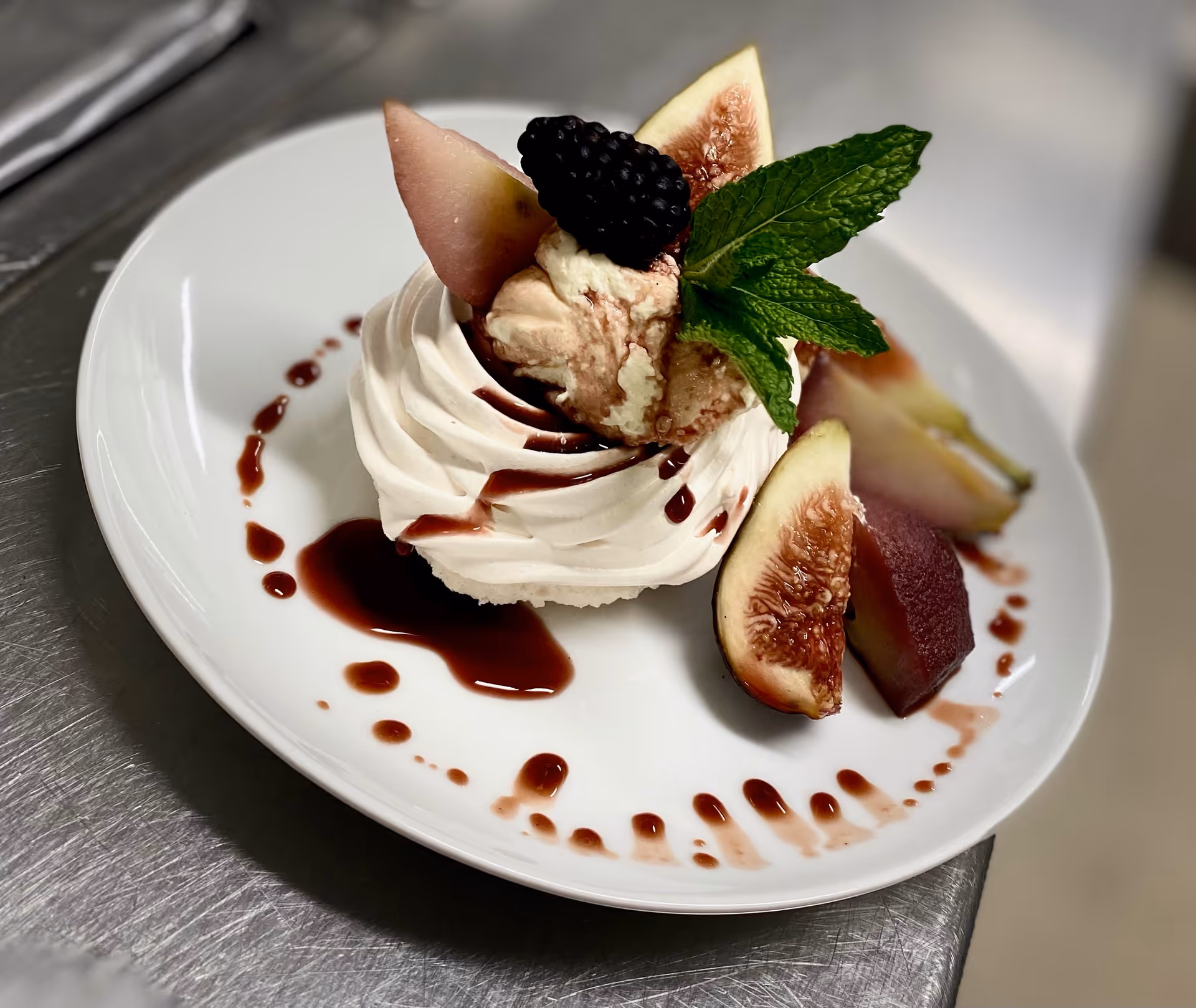 A plated dessert featuring a swirl of whipped cream or meringue topped with a blackberry, fresh figs, a mint sprig, and a drizzle of dark red sauce on a white plate.