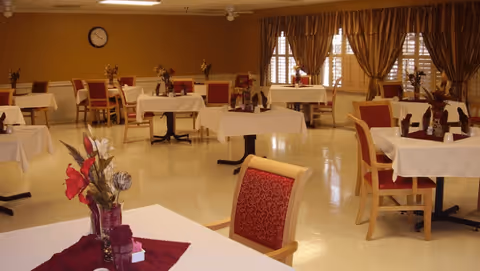 A dining room with multiple tables covered with white tablecloths and decorated with floral centerpieces. Each table is surrounded by wooden chairs with red cushioned seats and backs. The room has beige walls, large windows with brown curtains, and a clock on the wall.